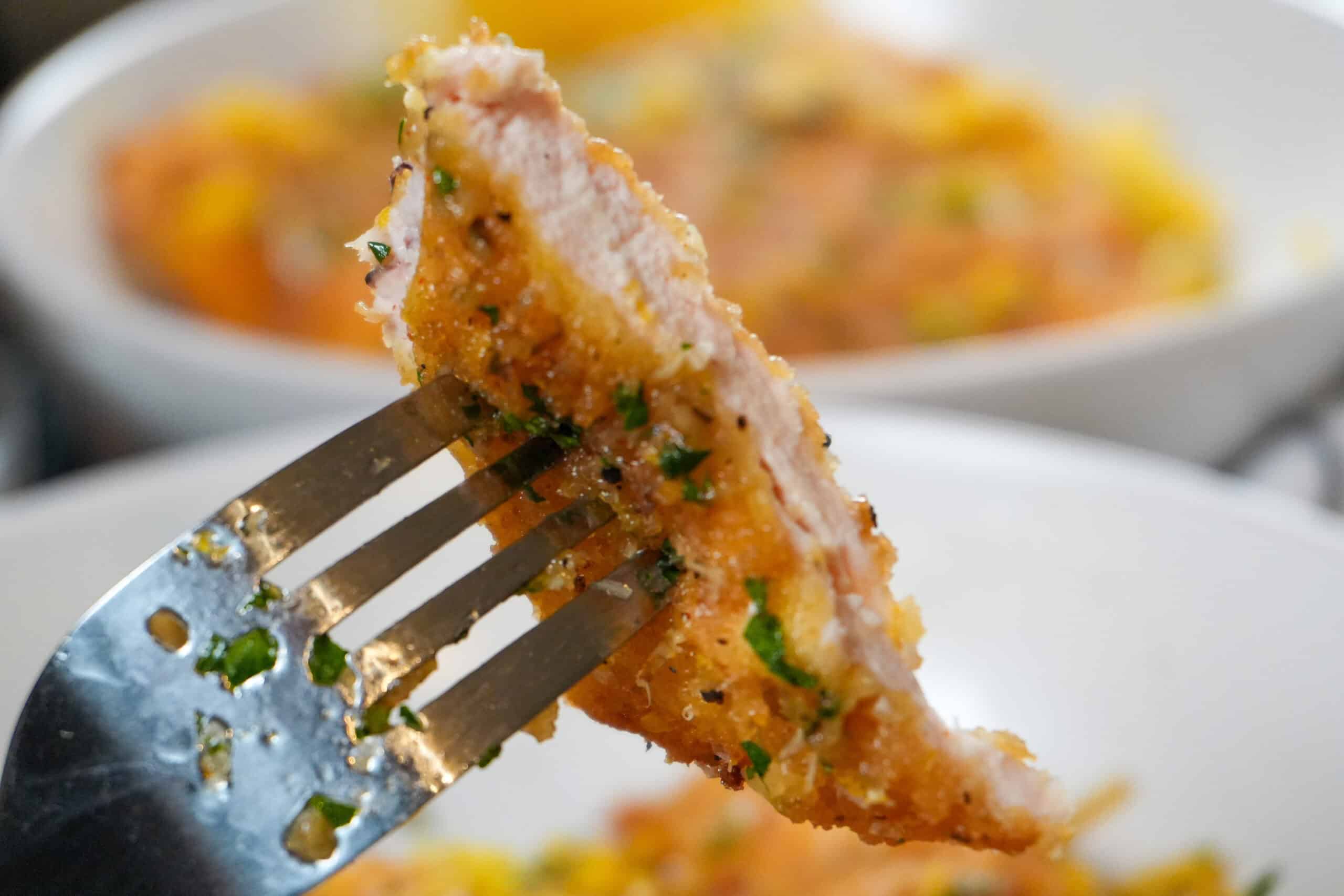 Crispy baked chicken on a fork, showcasing juicy meat and inviting side dishes in background.